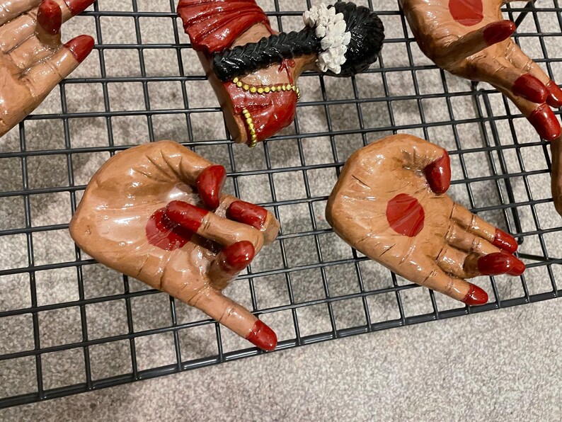 May include: Hand-shaped ceramic figurines with brown skin tones, red nail polish, and red dot accents. Some figurines have a red head covering and gold jewelry. The figurines are arranged on a black wire rack.