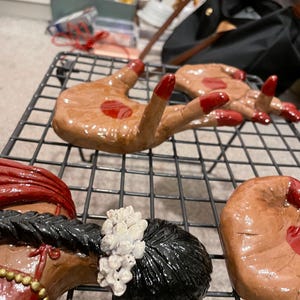 May include: Hand-shaped ceramic art pieces with red nail polish and a red drop of paint. The pieces are brown and are arranged on a black wire rack. Other ceramic pieces are also visible.