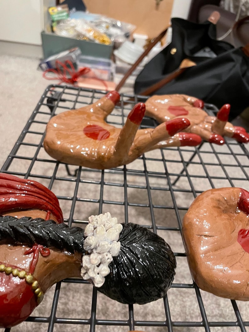 May include: Hand-shaped ceramic art pieces with red nail polish and a red drop of paint. The pieces are brown and are arranged on a black wire rack. Other ceramic pieces are also visible.