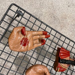 May include: A collection of hand-shaped figurines with red accents on the nails and palms. One figurine depicts a woman in a red and gold outfit with a black braid and white flower accents. The figurines are placed on a black wire rack.