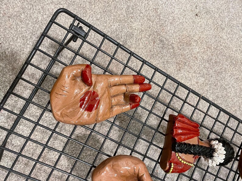 May include: A collection of hand-shaped figurines with red accents on the nails and palms. One figurine depicts a woman in a red and gold outfit with a black braid and white flower accents. The figurines are placed on a black wire rack.