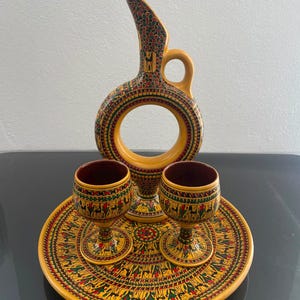 Puede incluir: Un juego de cerámica amarilla que incluye una jarra circular con asa, dos copas con interior oscuro y una bandeja redonda. Cada pieza está decorada con intrincados patrones repetitivos en tinta roja, verde y negra.