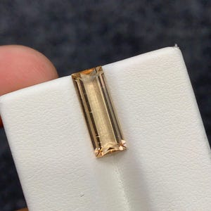 以下が含まれることがあります： 半透明のゴールデンブラウンの色合いを持つ、長方形のバゲットカットの宝石。石は光を反射する鋭くきれいなファセットを備えており、磨かれた表面と蜂蜜のような色合いを持つ、透明で細長い形状をしています。