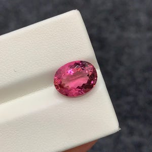 Può includere: Una pietra preziosa dal taglio ovale con un colore rosa acceso traslucido e una superficie sfaccettata che riflette la luce. La pietra è posizionata su un espositore bianco testurizzato, mettendone in risalto l'aspetto chiaro e lucido.