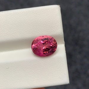 以下が含まれることがあります： 鮮やかなホットピンク色のオーバルカットの宝石。ファセット加工された表面が光を反射します。透明度が高く、内部に深みを感じさせるこの石は、質感のある白いディスプレイスタンドの上に置かれています。