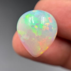 May include: A pear-shaped cabochon opal gemstone featuring a translucent white base with vibrant flashes of neon green, yellow, and orange play-of-color throughout the stone. The surface is smooth and polished, displaying a distinct teardrop silhouette.