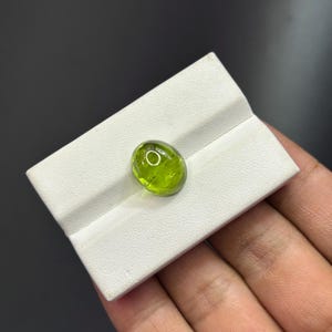 Op de afbeelding: Een doorschijnende, ovaalvormige peridoot edelsteen met een gladde, gepolijste cabochon slijpvorm. De steen heeft een levendige limoengroene kleur en is geplaatst in een witte displaystandaard met een horizontale gleuf.