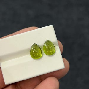 Op de afbeelding: Twee peervormige, doorschijnende limoengroene edelstenen met een glad, gepolijst oppervlak. De stenen worden naast elkaar getoond op een witte rechthoekige sieradenkaart, waarbij de levendige kleur en de druppelvorm zichtbaar zijn.