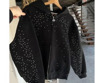 May include: Black zip-up hoodie with a constellation pattern of white star-shaped embellishments. The hoodie features a hood, front pockets, and a silver zipper. The garment is displayed on a wooden hanger.