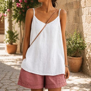 May include: A white camisole top with thin straps and a V-neck paired with dusty rose shorts. The outfit is made of a lightweight fabric, perfect for warm weather. A brown leather strap crosses the body.