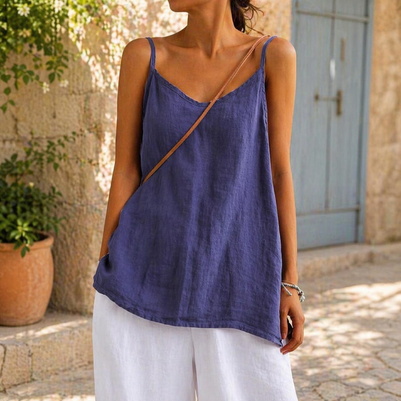 May include: A blue camisole top with thin straps and a V-neck. The top is paired with white pants. A brown strap crosses the body. The background includes a stone wall and a blue door.