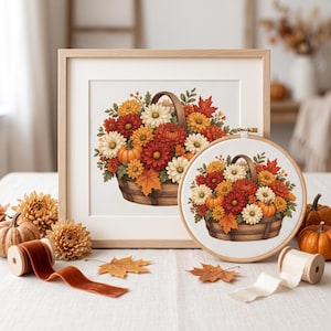 May include: A square framed embroidery and a circular embroidery hoop both featuring a stitched basket filled with autumn-colored chrysanthemums, maple leaves, and small pumpkins. The set includes spools of velvet ribbon and loose leaves.
