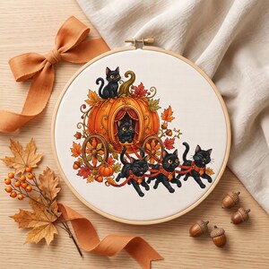 May include: A circular wooden embroidery hoop featuring a cross-stitch pattern of a pumpkin carriage drawn by three black cats, with a black cat perched on the pumpkin and another inside, decorated with orange and red autumn foliage.