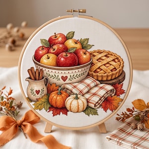 May include: A circular embroidery hoop featuring a printed design of a ceramic bowl filled with red and yellow apples, a lattice-crust pie, a small pitcher with cinnamon sticks, two pumpkins, a plaid cloth, and scattered autumn leaves.