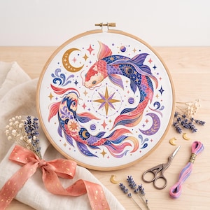 May include: A circular wooden embroidery hoop containing a white fabric panel with a cross-stitch design of two koi fish swimming in a circle around a central star. The fish feature pink, blue, and purple scales with celestial moon and star motifs.