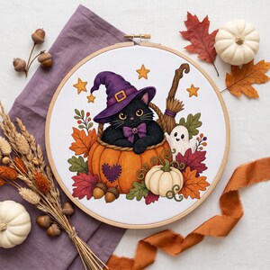May include: A cross-stitch embroidery in a round wooden hoop featuring a black cat wearing a purple witch hat and bow tie, sitting inside a large orange pumpkin. The design includes a small white ghost, autumn leaves, acorns, and yellow stars.