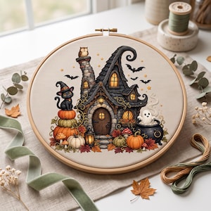 Könnte beinhalten: Ein runder Holzstickrahmen mit einem Kreuzstichmotiv, das ein Halloween-Häuschen mit Steinkamin, eine schwarze Katze mit Hexenhut auf Kürbissen, ein Gespenst in einem Kessel, Fledermäuse und Herbstblätter auf beigem Stoff zeigt.