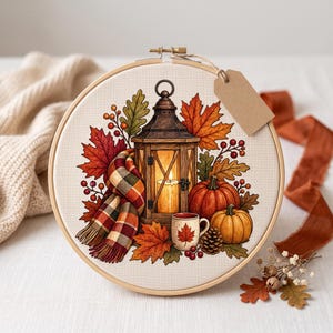 Peut inclure: Un tambour à broder rond en bois présentant un motif au point de croix composé d'une lanterne allumée, d'une écharpe à carreaux, de deux citrouilles, d'une tasse avec une feuille d'érable et de feuillage d'automne. Une étiquette en papier brun est attachée.