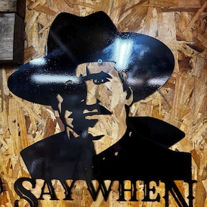 May include: Black metal wall art featuring a silhouette of a man in a cowboy hat. The artwork has the words "SAY WHEN" cut out at the bottom. The background is a wood composite.