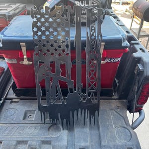 May include: A black metal wall art piece depicting the American flag with cut-out stars and stripes. The design incorporates oil derricks and rigs, with a textured, dripped effect at the bottom. The artwork is displayed on a truck bed.
