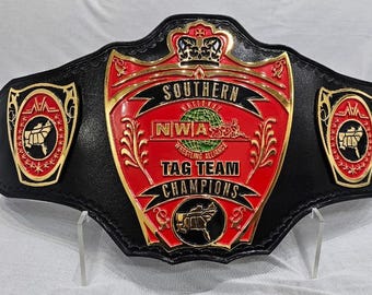 May include: A black wrestling championship belt with red and gold accents. The belt features the text "SOUTHERN NWA TAG TEAM CHAMPIONS" and a logo with a globe and a map of the southern United States.