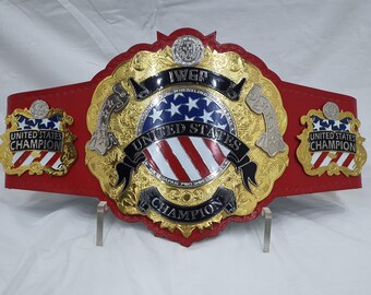May include: A gold and red wrestling championship belt with the words "United States Champion" and "IWGF" on it. The belt has a red leather strap and gold accents. The center of the belt features the American flag.