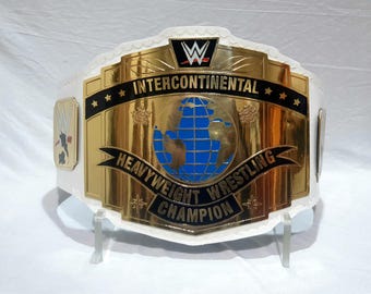 Puede incluir: Un cinturón de campeón de lucha libre de peso pesado intercontinental dorado y blanco. El cinturón presenta una placa central dorada con un diseño de globo azul y las palabras "Heavyweight Wrestling Champion". El cinturón tiene una correa blanca y el logo de la WWE.