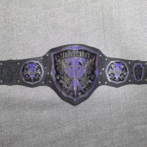 May include: A black and purple wrestling championship belt. The center plate features the word "Undertaker" in silver lettering, with a purple and silver emblem. The belt has silver studs and decorative accents.