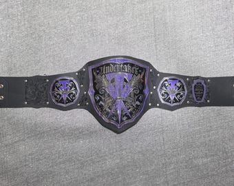 May include: A black and purple wrestling championship belt. The center plate features the word "Undertaker" in silver lettering, with a purple and silver emblem. The belt has silver studs and decorative accents.