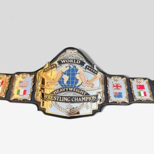 Könnte beinhalten: Ein gold-schwarzer World Heavyweight Wrestling Champion Gürtel. Der Gürtel hat ein großes zentrales Emblem mit einem Globus, Adlern und den Worten "World Heavyweight Wrestling Champion". Kleinere Embleme mit Flaggen befinden sich auf jeder Seite.