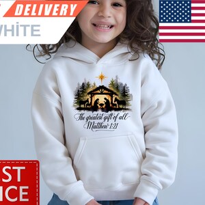 May include: White hooded sweatshirt featuring a Nativity scene graphic with the text "The greatest gift of all Matthew 1:21." The graphic includes silhouettes of figures, trees, and a star. The sweatshirt has a front pocket and is being worn by a person.