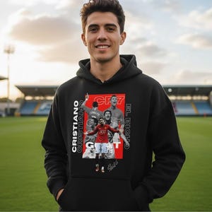 May include: Black hoodie featuring a collage of images of Cristiano Ronaldo, with the text "CR7" and "Cristiano" in white. The hoodie has a drawstring hood and a front pocket. The background shows a soccer field.