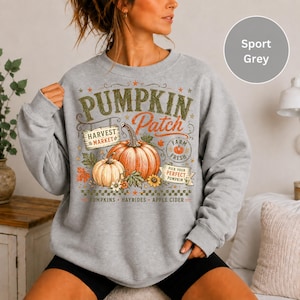 May include: A sport grey crewneck sweatshirt featuring a graphic print with the text PUMPKIN PATCH, HARVEST MARKET, FARM FRESH, and PUMPKINS HAYRIDES APPLE CIDER. The design includes orange pumpkins, green leaves, sunflowers, and a checkered border.