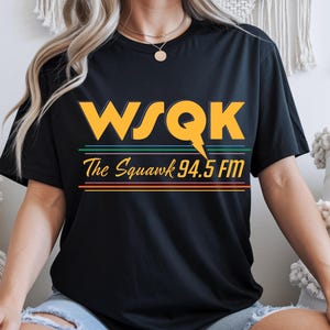 May include: Black t-shirt with the text "WSQK The Squawk 94.5 FM" in yellow, green, pink, and orange. The design features a lightning bolt through the letter "Q". The shirt is a casual wear item.