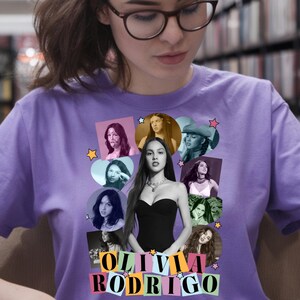May include: A lilac-colored t-shirt featuring a collage of images of Olivia Rodrigo. The design includes various photos of the singer, surrounded by star shapes, and the name "OLIVIA RODRIGO" in colorful block letters.