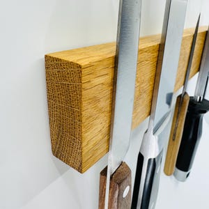 Può includere: Una striscia magnetica rettangolare in legno di rovere chiaro montata orizzontalmente. Il supporto sostiene quattro coltelli da cucina con vari design di manico, inclusi legno e materiali sintetici neri, con le lame in metallo visibili.