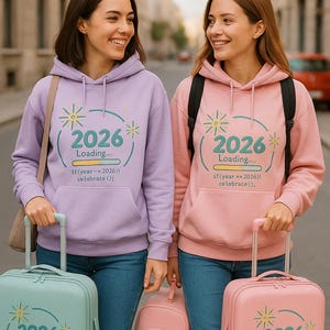 Peut inclure: Deux personnes portant des sweats &agrave; capuche assortis avec le texte "2026 Loading" et des bagages. Les sweats &agrave; capuche sont de couleur lilas et rose. L'une a une valise bleu clair, l'autre une valise et un sac &agrave; dos roses. Le texte comprend la phrase "if (year == 2026)! celebrate();".