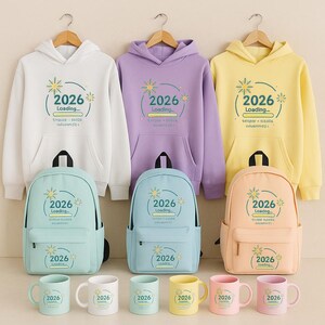 Peut inclure: Ensemble de sweats &agrave; capuche, sacs &agrave; dos et mugs aux couleurs pastel assorties. Les sweats &agrave; capuche sont blancs, lavande et jaunes. Les sacs &agrave; dos et les mugs sont bleu clair, p&ecirc;che, jaune et rose. Chaque article porte l'inscription "2026 Loading..."