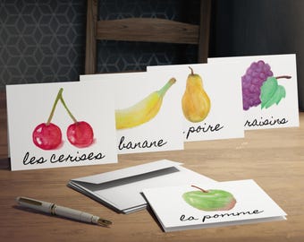 May include: Greeting cards with watercolour fruit illustrations: cherries, banana, pear, grapes, and an apple. Each card has the fruit's name in cursive. A pen and envelopes are on a wooden surface.