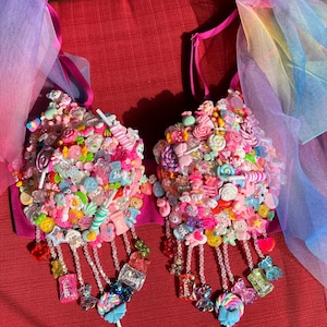 May include: A pink bra covered in colorful candy-themed embellishments, including lollipops, gummy bears, and other sweets. The bra has beaded fringe and is adorned with sheer, rainbow-colored fabric.
