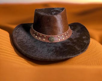 May include: A brown cowboy hat with a dark brown brim and crown. The hat has a brown leather band with small, gold-coloured studs and a circular emblem. The hat is on a sandy surface.