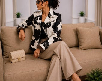 May include: A black and white cow-print leather jacket with a notched collar, silver-tone metal zippers, and a buckled cuff detail. The jacket features a dark brown interior lining, paired with wide-leg beige trousers and cream-colored pumps.