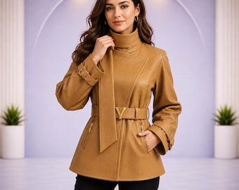May include: A tan jacket with a high collar, long sleeves featuring buckled cuffs, and a waist belt with a gold-toned V-shaped buckle. The jacket includes a vertical front panel detail, two zippered side pockets, and is paired with black trousers.