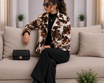 May include: A brown and white cow-print moto jacket with a wide collar, long sleeves, and metallic zippers. The jacket is worn over a black mock-neck top and black trousers, paired with black pointed-toe boots and a black quilted shoulder bag.