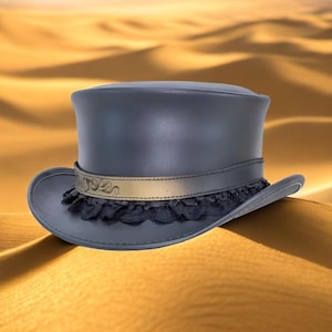 Puede incluir: Un sombrero de copa gris oscuro con una banda dorada y ribete de encaje negro. El sombrero tiene una corona alta clásica y un ala ancha. La banda dorada presenta un diseño ornamentado. El sombrero está sobre un fondo borroso del desierto.