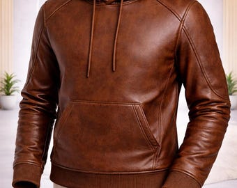 May include: A brown leather hoodie featuring a front kangaroo pocket, long sleeves with ribbed cuffs, a matching ribbed hem, and a hood with integrated brown drawstrings. The garment displays a smooth texture and visible stitching along the shoulders.