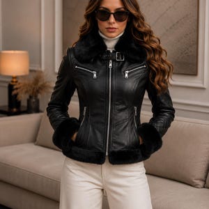 May include: A black leather jacket featuring a plush black faux fur collar and cuffs, a silver front zipper, two zippered chest pockets, two lower zippered pockets, and a buckled strap at the neck, worn over a white turtleneck sweater.