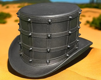 May include: A black leather top hat with a wide brim. The hat features a series of studded straps that encircle the crown. The hat is sitting on a sandy surface.