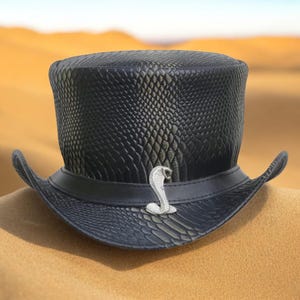 Puede incluir: Un sombrero de copa negro con textura de piel de serpiente y un emblema de cobra plateada. El sombrero tiene un ala ancha y una banda de cuero. Un accesorio único.