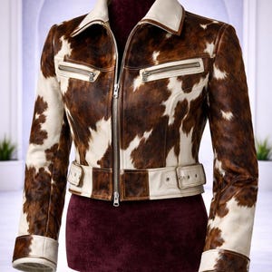 May include: A cropped jacket featuring a brown and white cowhide print pattern. The jacket includes a solid white collar, a central silver zipper, two horizontal zippered chest pockets, and a white waist belt with a buckle closure.